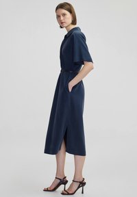 Abito blu navy con colletto, maniche corte e vita regolabile con cintura. Presenta tasche laterali e uno spacco frontale. Abbinato a sandali neri con tacco.