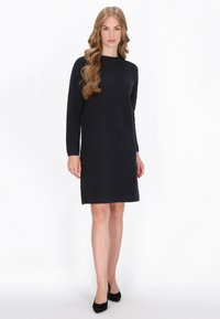 Robe en maille bleu marine à manches longues, col rond et paillettes discrètes. Coupe droite arrivant au genou. Talons noirs.