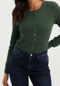 Mörkgrön strukturerad cardigan med rund halsringning, knappstängning fram och långärmar. I kombination med mörk denimjeans.