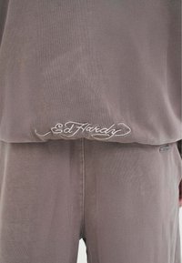 Tuta in velour grigio con logo "Ed Hardy" ricamato. Texture liscia, vestibilità rilassata, con una leggera lucentezza che mette in risalto il materiale.
