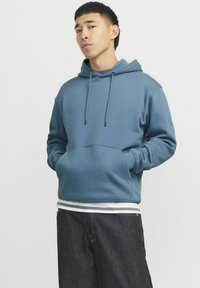Blå hættetrøje lavet af blødt stof, med en frontlomme, justerbare snore og ribbede manchetter. Sammen med mørke denimbukser.