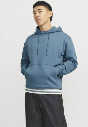 Blue hoodie made of soft fabric, featuring a front pocket, adjustable drawstrings, and ribbed cuffs. Paired with dark denim pants.