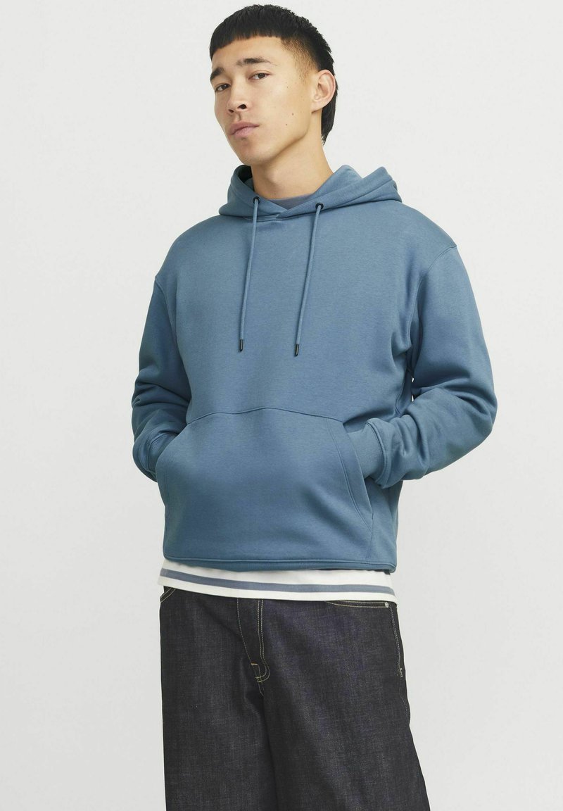 Haut à capuche bleu en tissu doux, avec une poche frontale, des cordons de serrage ajustables et des poignets côtelés. Associé à un pantalon en denim foncé.