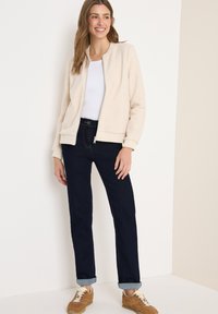 Mujer sonriendo, vistiendo una chaqueta de forro polar cremosa con cremallera, camiseta blanca, jeans oscuros de puño y zapatillas color beige, de pie contra una pared de color claro.