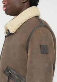 Chaqueta de cuero marrón con un grueso cuello de borreguillo crema, bolsillo con cremallera lateral y un sutil parche de marca en la manga.