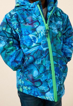 Enfant portant une veste à capuche bleue avec une fermeture éclair verte et un motif graffiti de dinosaure, les mains dans les poches.
