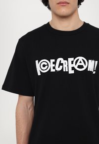 Melna kokvilnas T-krekls ar izteiksmīgu balto tekstu "SALIA" un apļveida logotipu ar simbolu "A". Brīvs piegriezums un īsas piedurknes.