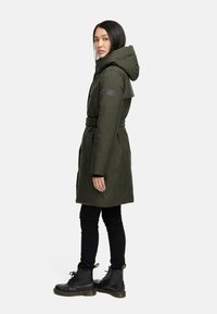 Oliven grøn hætteparka med bælte, struktureret stof og en sidelomme med lynlås. Sammen med sorte skinny jeans og sorte snørebåndsstøvler.