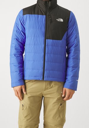 Homme portant une veste matelassée North Face bleue et noire et un pantalon cargo beige avec des poches zippées, debout devant un fond uni.