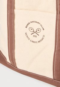 Saco de canvas em creme e castanho. Apresenta um logotipo com raquetes cruzadas e o texto "Borg Racquet Club" e "Monte Carlo, Mónaco." Detalhes em costura.