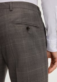 Grijze geblokte broek met een achterzak met knoopsluiting, getailleerde pasvorm en subtiele textuurcontrast. Witte shirt zichtbaar bij de kraag.