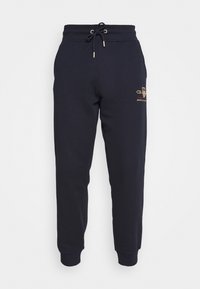 Námořnické modré tepláky vyrobené z měkké tkaniny, s elastickým pasem a šňůrkou, a zlatým logem umístěným na boku.