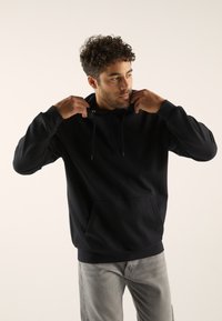 Sudadera negra con capucha, bolsillo canguro, cordones y puños acanalados. Combinada con pantalones grises sobre un fondo neutro.