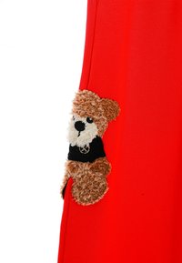 Prenda roja con un apliqué texturizado de oso de pelo marrón y una camisa negra, posicionada cerca de la costura lateral.