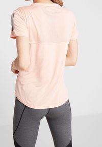 Trenérové tričko v broskvové barvě s síťovanou horní částí, krátkými rukávy a zaobleným lemem, v kombinaci se šedými legínami s černými akcenty.