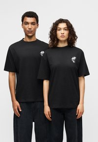 Μαύρα βαμβακερά T-shirt με κοντά μανίκια, διακοσμημένα με λευκό λουλουδάτο σχέδιο στο αριστερό στήθος. Συνδυασμένα με άνετο μαύρο τζιν παντελόνι.