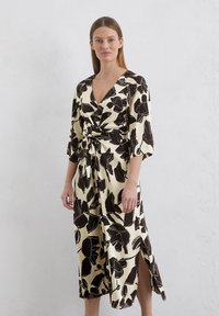Wrap dress in cream fabric with black floral pattern, V-neck, short sleeves, and side slit. Smooth texture, fitted waist with twist detail.