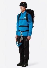 Chaqueta de forro polar azul con cremalleras, arnés de escalada negro equipado con equipo de escalada, pantalones y botas negras, y un casco negro.