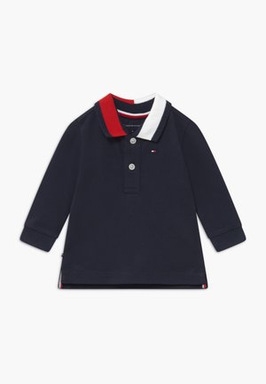 Navyblå langærmet polo med rød og hvid krave, treknapslukning og sideslidser. Har mærkelogo på brystet. Fremstillet i bomuld.