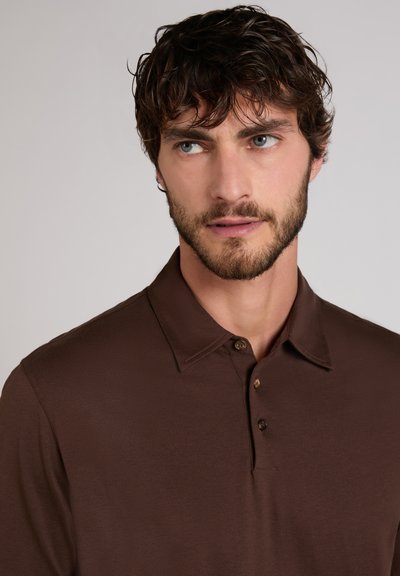 Mann mit lockigem braunem Haar und Bart, der ein dunkelbraunes, geknöpftes Poloshirt trägt und leicht seitlich vor einem einfarbigen Hintergrund blickt.