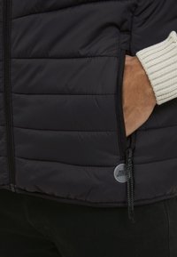 Schwarze Puffjacke mit horizontalem Steppmuster, seitlicher Reißverschlusstasche und hellen Rippbündchen. Mit einem Logo am Reißverschlussgriff.