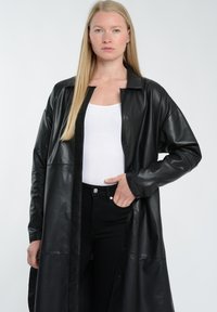 Lang sort læderkåbe med kravesnit, knaplukning og brede ærmer. Matchet med en hvid top og sorte jeans. Glat tekstur.