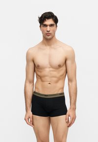 Homme torse nu aux cheveux foncés, debout face à l’avant, portant un boxer noir avec une ceinture olive portant l’inscription « BOSS » sur un fond gris uni.