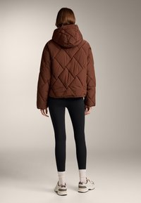 Brun quiltet pufferjakke med hætte, der har et diamantmønster. Bæres over sorte leggings, med synlige hvide sokker og sko.