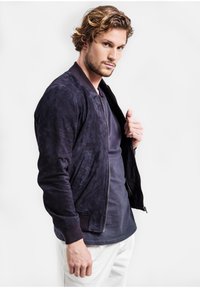 Námornícky zamatu bombera s rebrovanými manžetami a golierom, so zipsom a bočnými vreckami. Nosený na tmavom tričku a bielych nohaviciach.