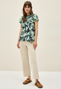 Blusenshirt mit floralem Muster in Grüntönen auf dunklem Hintergrund; kombiniert mit hellbeigen weiten Hosen und braunen Clogs.