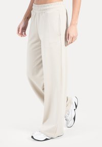 Beige bukser med vide ben lavet af blødt stof med elastik i taljen. Har sidelommer og et diskret logo broderet foran.