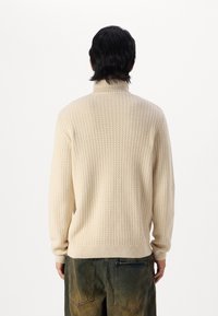 Hellbeige Rollkragenpullover mit strukturiertem, geripptem Muster. Gerippte Bündchen und Saum. Getragen mit weit geschnittenen, dunkelgrünen Hosen.