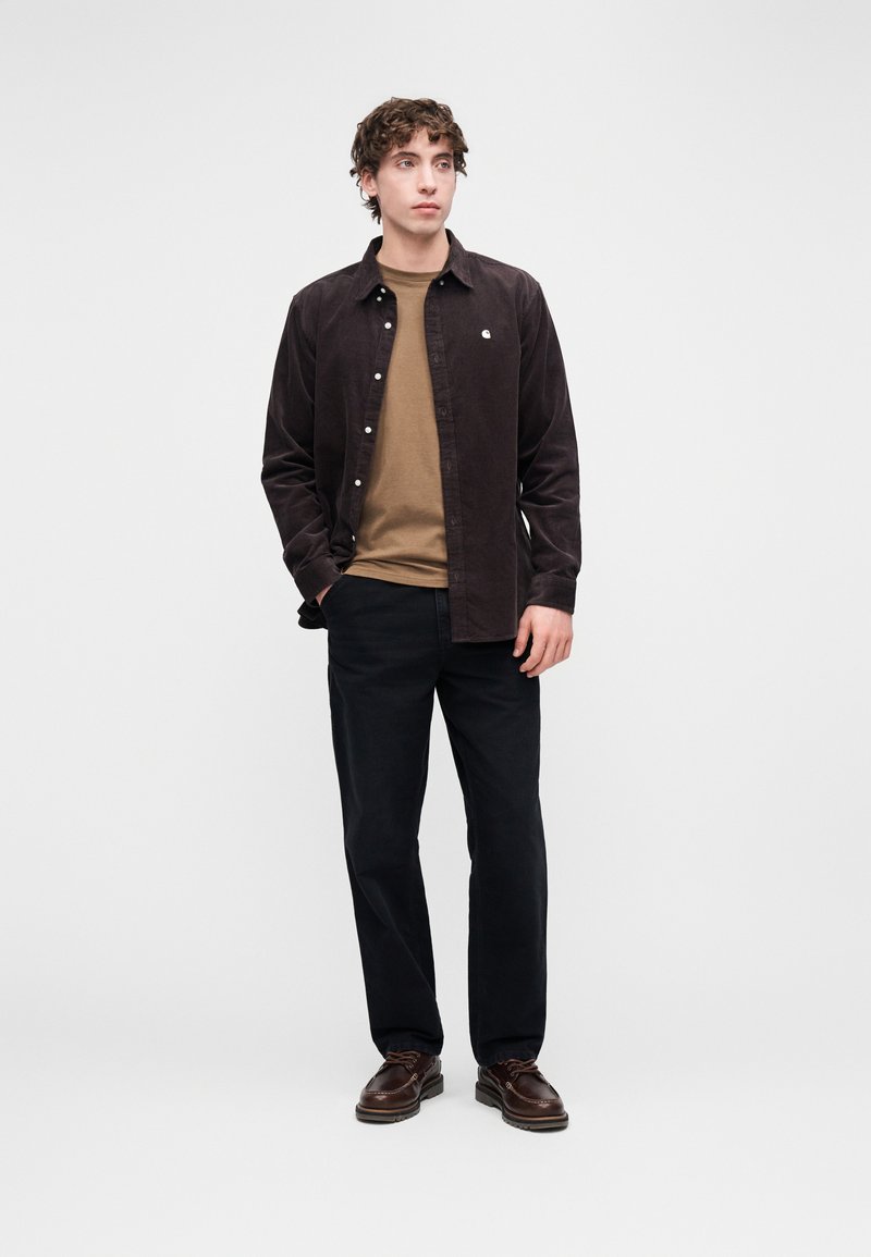 Camicia marrone di velluto a coste con bottoni sopra una t-shirt beige, pantaloni neri e scarpe marroni. La camicia presenta un piccolo logo e bottoni.