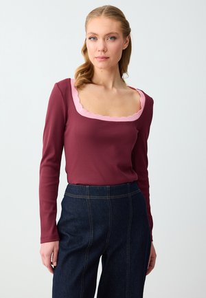 Vrouw draagt een bordeauxrode top met lange mouwen en een roze, geschulpte vierkante halslijn, samen met donkerblauwe hoge taille broek, staand tegen een witte achtergrond.