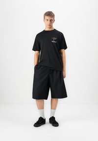 Ung mann iført svart T-skjorte med gult kengurulogos og tekst som sier "Australia", svarte vide shorts, hvite sokker og svarte joggesko.