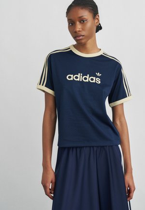 Mladi žena nosi tamnoplavu Adidas majicu s bež obrubom i logotipom, uparen s tamnoplavim donjim dijelom, na jednobojnoj svijetlosivoj pozadini.