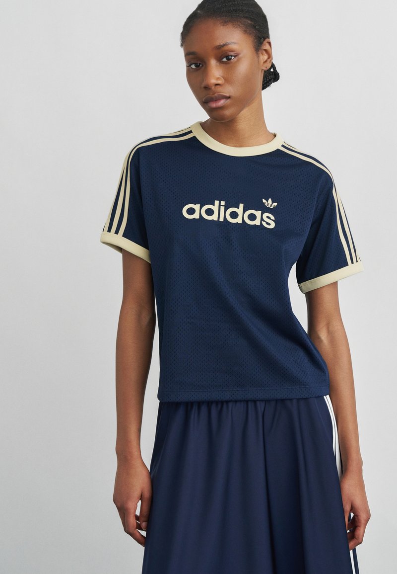 Mladá žena ve tmavě modrém tričku Adidas s béžovým lemováním a logem, kombinovaném s tmavě modrými kalhotami na jednobarevném světle šedém pozadí.