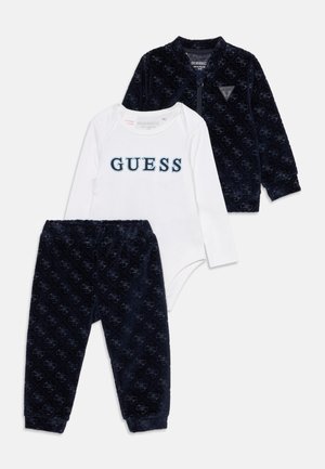 Ensemble de vêtements bébé trois pièces avec veste bleu marine à motifs, pantalon assorti et body blanc à manches longues avec logo "GUESS".