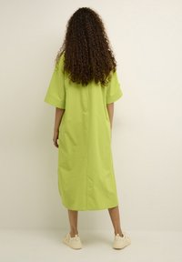 Abito di cotone oversize verde lime con maniche corte, lungo fino al ginocchio. Presenta una cucitura centrale sul retro, abbinato a sneakers di colore chiaro.