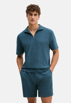 Hombre con cabello rizado que lleva un pulóver de medio cierre de manga corta en color verde azulado y pantalones cortos a juego, de pie con las manos en los bolsillos.