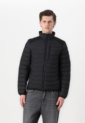 Zwarte pufferjack met horizontaal gestikt patroon, hoge kraag en ritssluiting. Bevat een borstzak en een gladde textuur. Gecombineerd met grijze jeans.