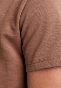 Hnědý přiléhavý tričko s hladkým povrchem, krátkým rukávem a jemně vroubkovaným výstřihem. Spodní lem má úhledné švy.