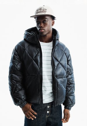 Junger Mann, der eine schwarze gesteppte Jacke, ein weiß gestreiftes Hemd, dunkle Jeans und eine weiße Kappe mit dem Text "Manhattan" trägt, steht vor einem weißen Hintergrund.