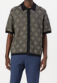 Mann trägt ein kurzärmliges, geknöpftes schwarz-beige gestreiftes Hemd mit schwarzem Kragen, kombiniert mit blauen Hosen vor einem schlichten Hintergrund.