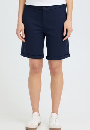 Jambes d'une personne portant un short bleu marine avec revers, des baskets blanches et un haut blanc, debout devant un fond clair uni.