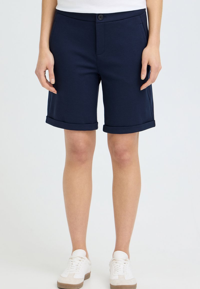 Jambes d'une personne portant un short bleu marine avec revers, des baskets blanches et un haut blanc, debout devant un fond clair uni.