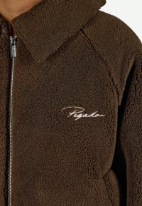 Giacca in pile marrone con chiusura a zip, caratterizzata da un colletto alto e un logo ricamato "Pegadon" in bianco sul petto. Texture morbida.
