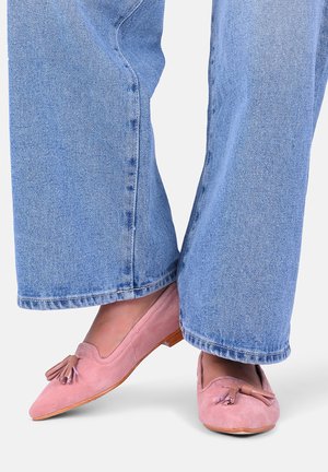 Person iført lyseblå jeans med vide ben og pink ruskind slip-on loafers med kvastdetaljer på en hvid baggrund.