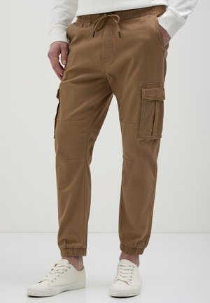 Uomo con pantaloni cargo marroni con elastici alle caviglie e scarpe da ginnastica bianche con lacci, in piedi con le mani nelle tasche.