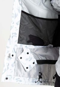 Let jakke med et hvidt og gråt camouflagemønster, der har en meshlomme, trykknapper og elastisk snor i kanten.